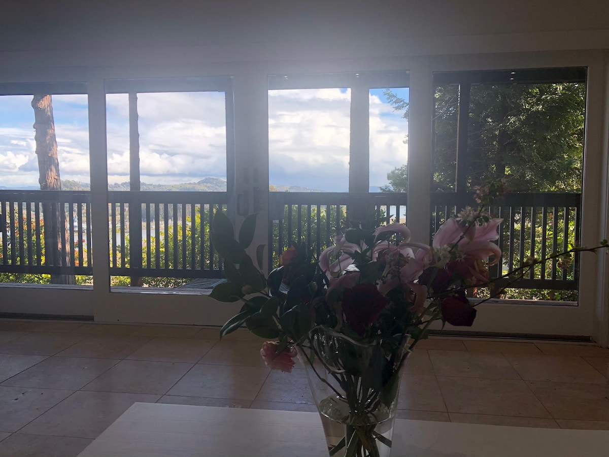Large windows provide a stunning view of Richardson Bay and the surrounding landscape. A vase filled with colorful flowers rests on a light-colored table, adding a touch of nature indoors. The serene atmosphere is complemented by the natural light streaming into the space.