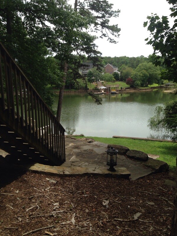 A stone pathway leads down to the water's edge, surrounded by lush greenery and mature trees. A lantern sits on the ground, providing soft illumination. Across the calm lake, residential structures are visible, integrated within the natural landscape.