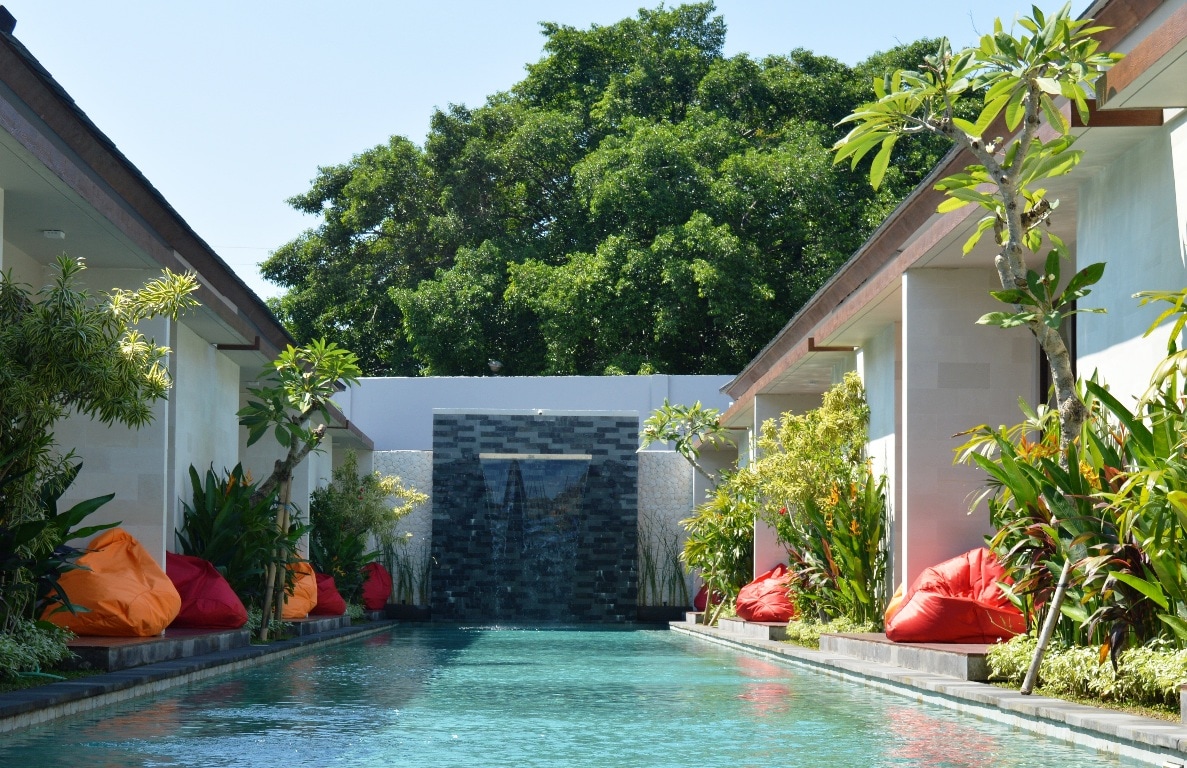 A peaceful pool area is surrounded by lush greenery and flowering plants. Colorful bean bags are positioned along the poolside, providing spots for relaxation. A water feature at the far end adds a calming touch to the serene environment.