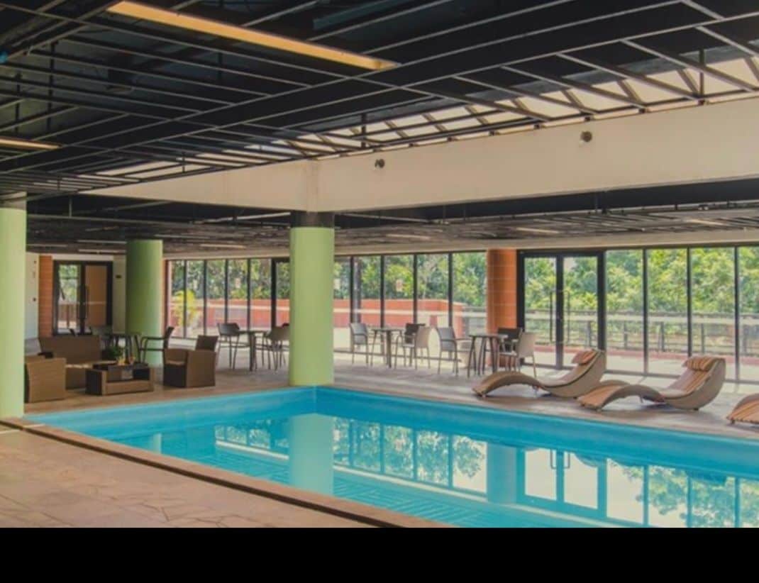 An indoor pool area is visible, framed by large windows that invite natural light. Comfortable seating arrangements are positioned on one side, while lounge chairs are placed beside the pool. A patio area is discernible in the background, contributing to the relaxing atmosphere.