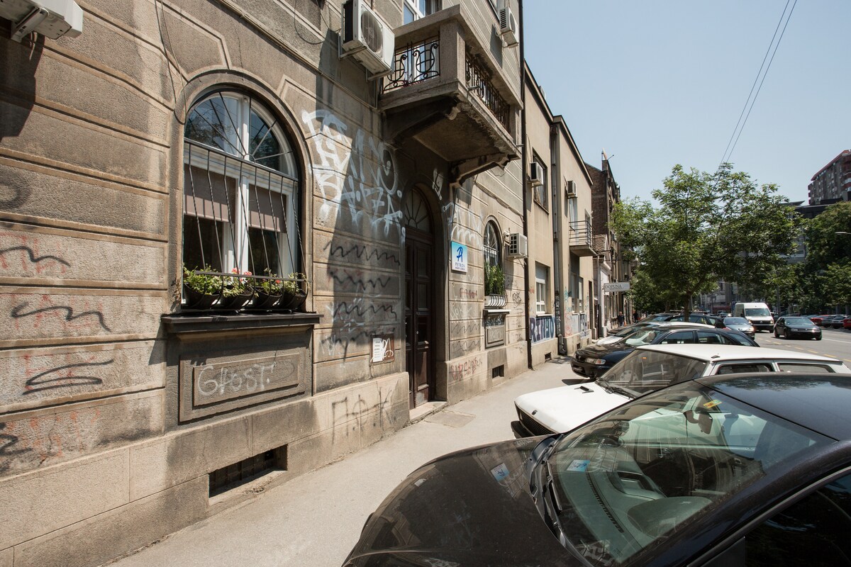 A street view showcases a historic building with arched windows and decorative stonework. The entrance is framed by a balcony adorned with flower pots. Nearby vehicles are parked along the road, and multiple trees provide greenery in the background.