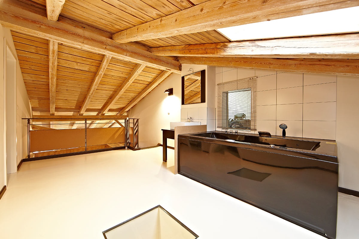 An open loft space showcases a sleek black bathtub and modern vanity against a backdrop of warm wooden beams. Natural light filters through skylights, illuminating the area, while a railing surrounds the upper level, creating a spacious and inviting atmosphere.