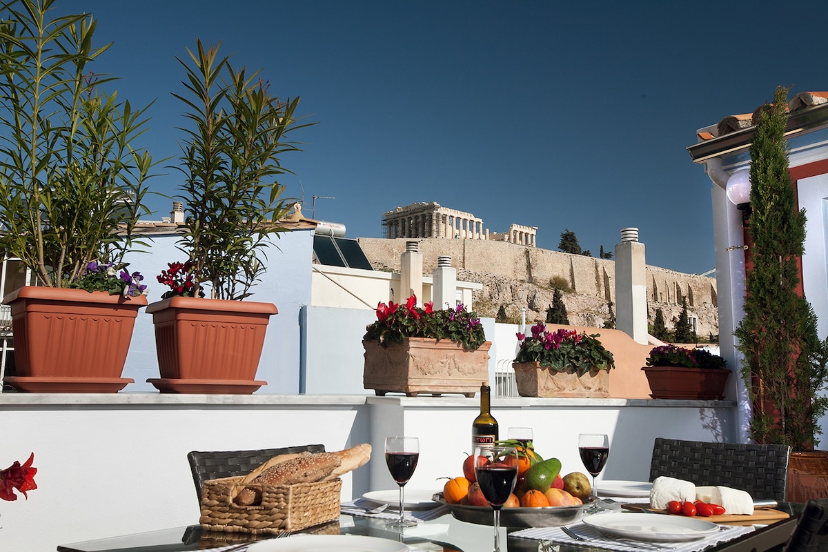A private roof garden terrace features a dining table set for meals, surrounded by vibrant potted plants and a view of the Acropolis. Fresh fruits and a basket of bread are arranged on the table, accompanied by bottles of wine. The blue sky enhances the serene atmosphere.