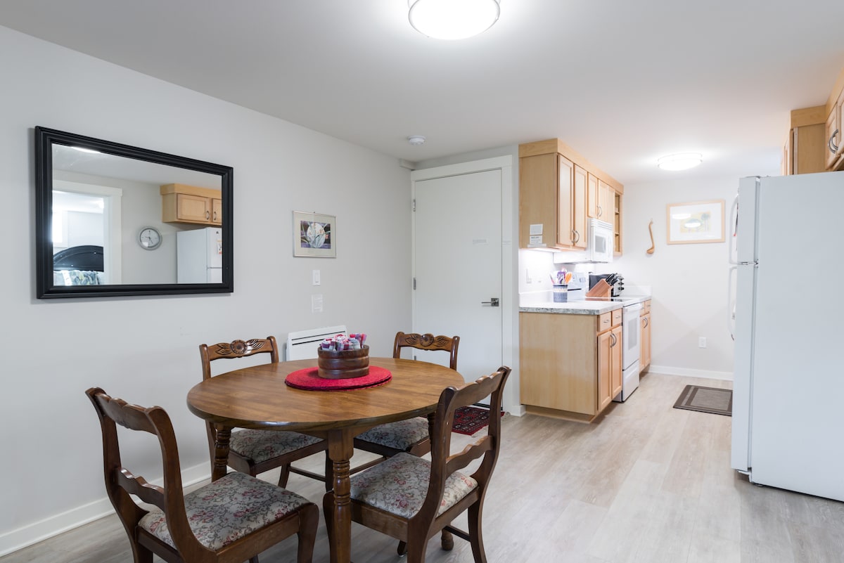 The kitchen area features a round wooden dining table with four chairs, accentuated by a red placemat. Light wood cabinetry complements the modern appliances, including a refrigerator and stove. A large mirror and wall art add visual interest to the bright, neutral-toned space.