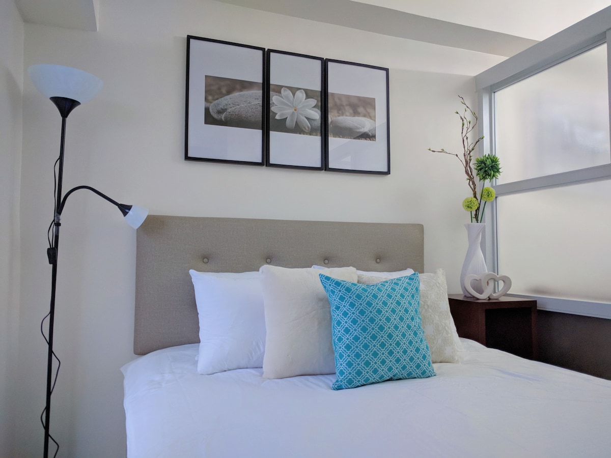 The bedroom features a queen-sized bed adorned with white and textured pillows. Above the headboard, three framed images of stones and flowers provide a calming visual. A stylish lamp and a decorative vase with greenery sit nearby, enhancing the space's simplicity.