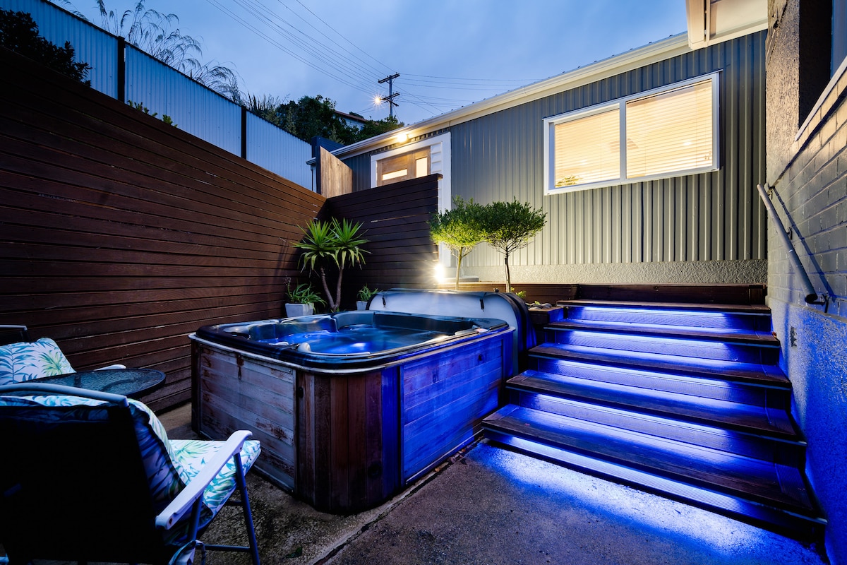 A private spa pool is situated beside a well-lit walkway, illuminated with blue lighting. The space is flanked by wooden fencing and features a small garden area with plants. Chairs are positioned for relaxation, inviting guests to enjoy the tranquil setting.
