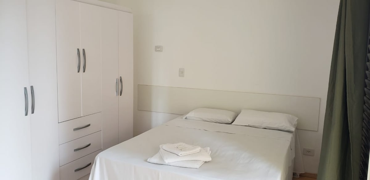A well-lit bedroom features a spacious bed neatly made with white linens and pillows. A multi-drawer wardrobe stands against the wall, providing ample storage space. Soft green curtains frame the window, allowing natural light to filter into the room.