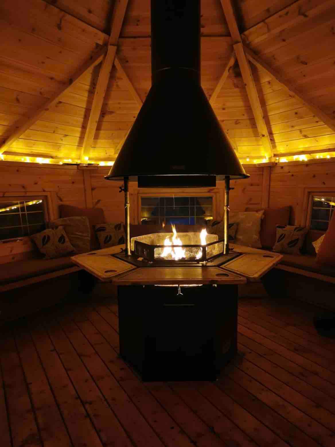 The interior of a cozy Norwegian BBQ cabin features a central BBQ pit surrounded by wooden seating. Warm light from string lights illuminates the walls, creating an inviting atmosphere. The cabin is structured in a hexagon shape, enhancing the communal feel for social gatherings.