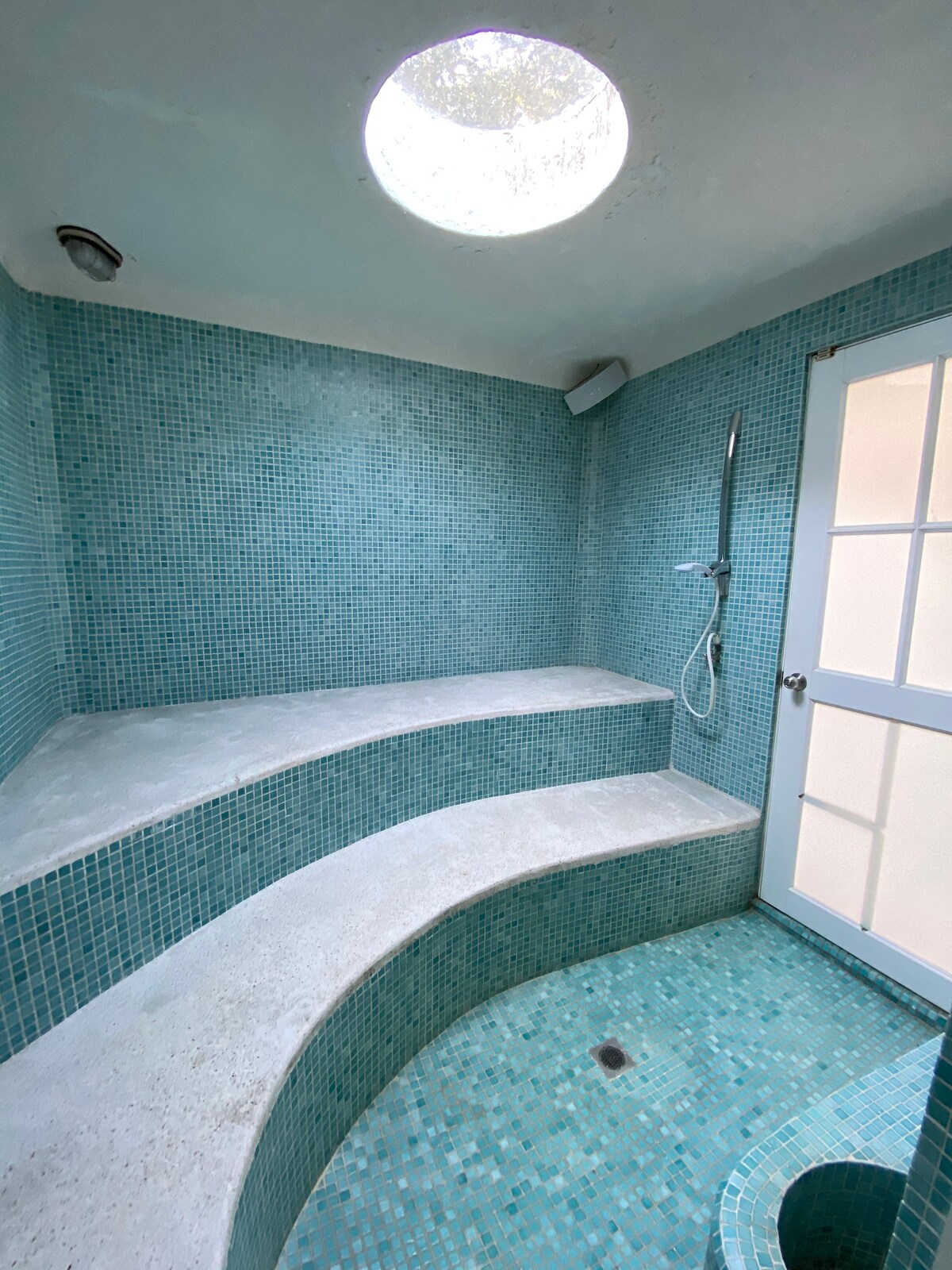 A tranquil steam bath area is highlighted with turquoise mosaic tiles covering the walls and curved seating. A showerhead is mounted on the wall, and a glass door allows natural light to enter, enhancing the calming ambiance of the space.