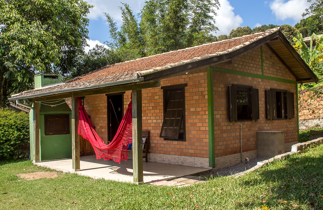 A charming brick cabin is set amidst a lush green landscape. A shaded porch features a vibrant red hammock, inviting relaxation. The exterior showcases wooden accents and is surrounded by well-maintained grass and trees, creating a peaceful outdoor setting.