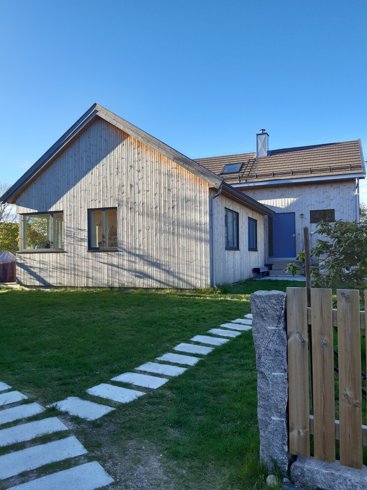 A modern wooden house is presented with a sloped roof and a combination of large windows on the façade. A pathway of stone pavers leads towards the entrance, surrounded by lush green grass and minimal landscaping.