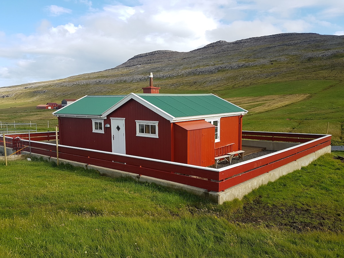 A red cottage is set in a grassy landscape, featuring a green roof and white trim. Surrounding the cottage is a low fenced area with a wooden table and benches. Mountains rise softly in the background under a partly cloudy sky.