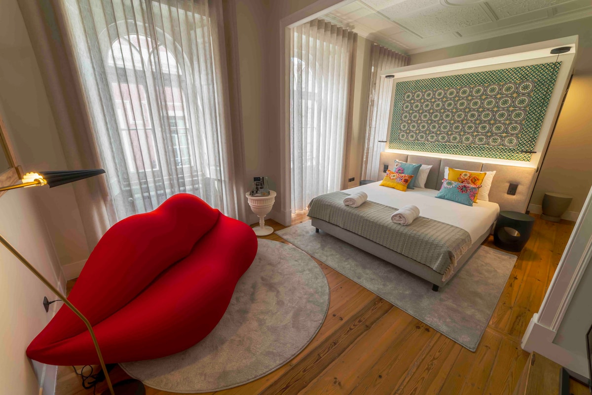 A contemporary room features a spacious bed with neutral bedding and colorful decorative pillows. A striking red lip-shaped chair adds a playful touch, while soft light filters through sheer curtains. Wooden flooring complements the overall design, enhancing the welcoming environment.