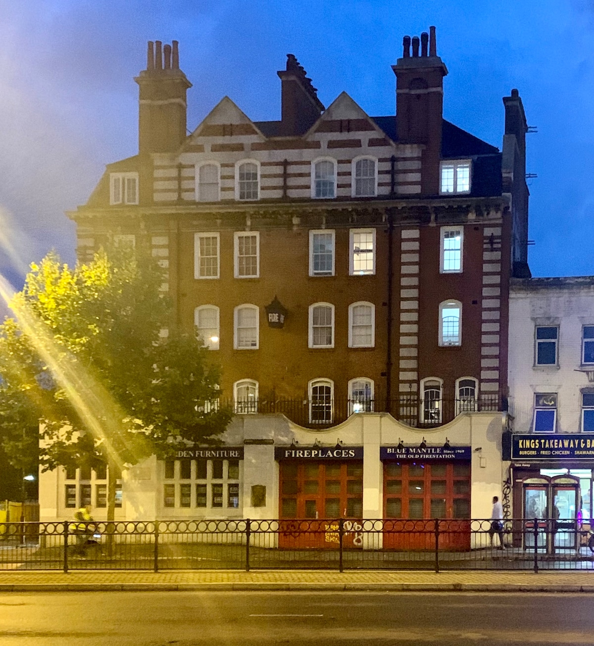 A historic building is seen at dusk, featuring a mix of red bricks and white accents. Its architectural details include prominent chimneys and a series of windows that reflect the evening light. The ground level contains commercial signage, enhancing the urban feel of the scene.