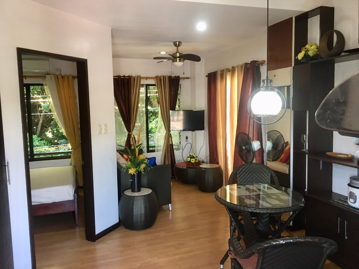 An open living area is featured, showcasing a modern design with warm tones. A round dining table sits centrally, surrounded by rattan chairs. A small television is visible, and large windows allow natural light to enter, framed by colorful curtains. A cozy sleeping area is seen through a doorway.