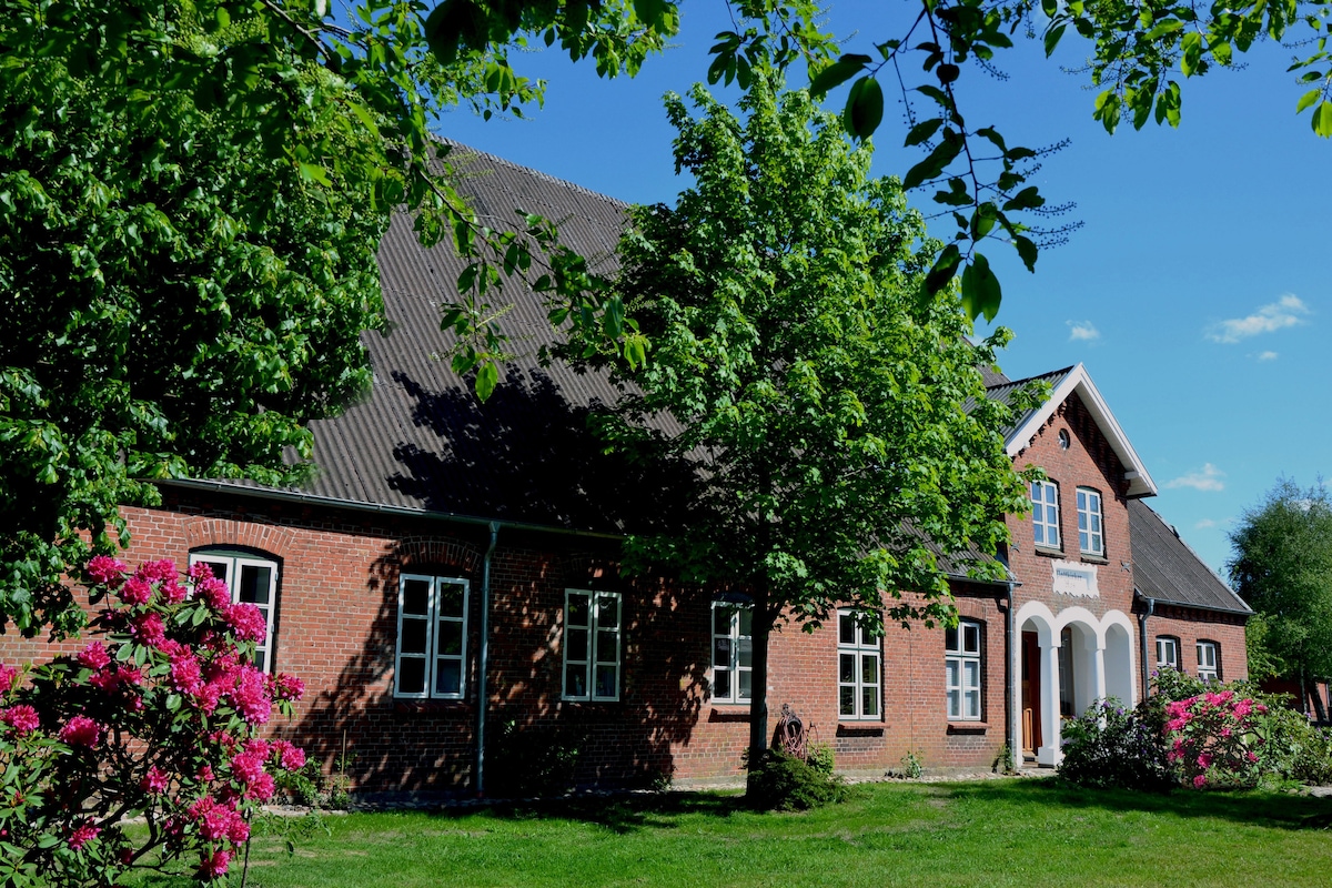 A charming old farmhouse is surrounded by lush greenery and blooming flowers. The red brick facade is complemented by white-framed windows and a welcoming entrance. Bright sunlight bathes the exterior, highlighting the natural beauty of the landscape.