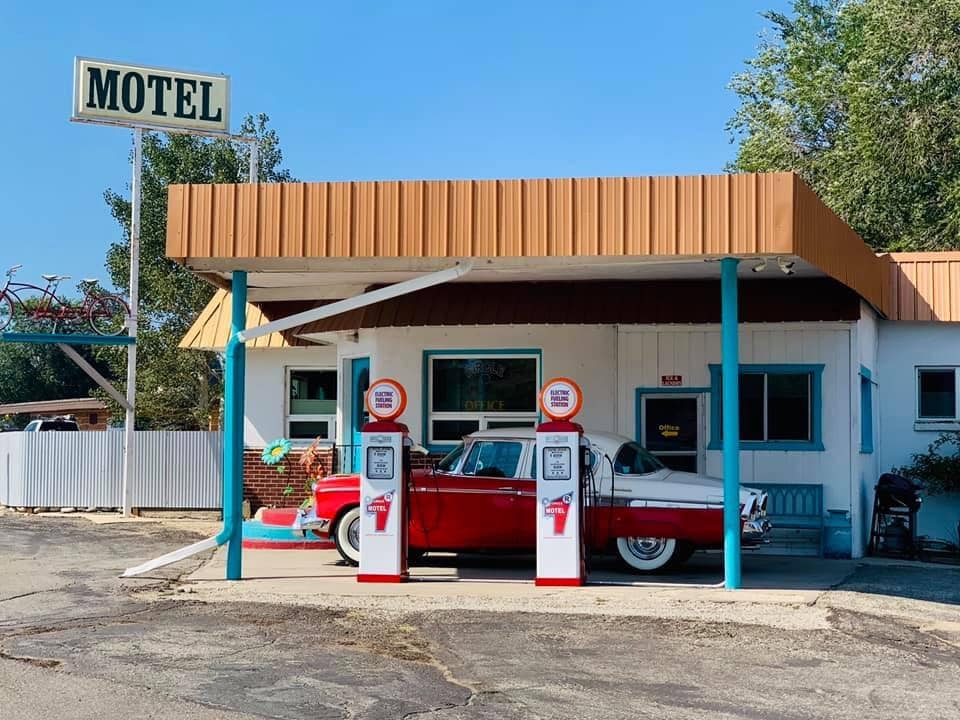 The exterior of a retro-inspired motel features a vintage car parked in front, flanked by two classic gas pumps. The building showcases a mix of modern and nostalgic design elements, with a bright orange awning and blue window frames, set against a clear sky.