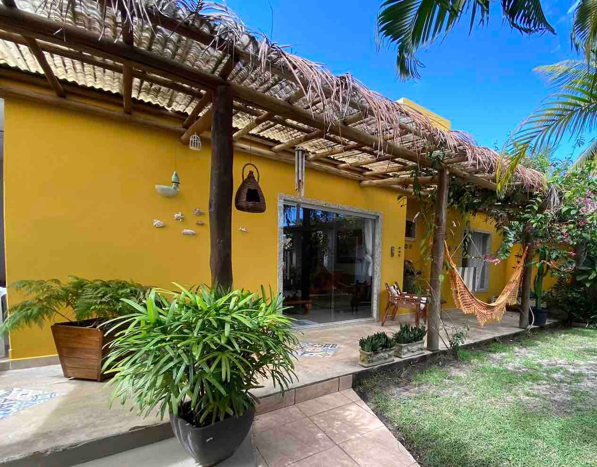 A bright yellow exterior is highlighted by a thatched awning and a cozy hammock. Lush greenery is present in large pots, contributing to the inviting outdoor space. Large glass doors provide a view into the interior, enhancing the connection between indoors and outdoors.