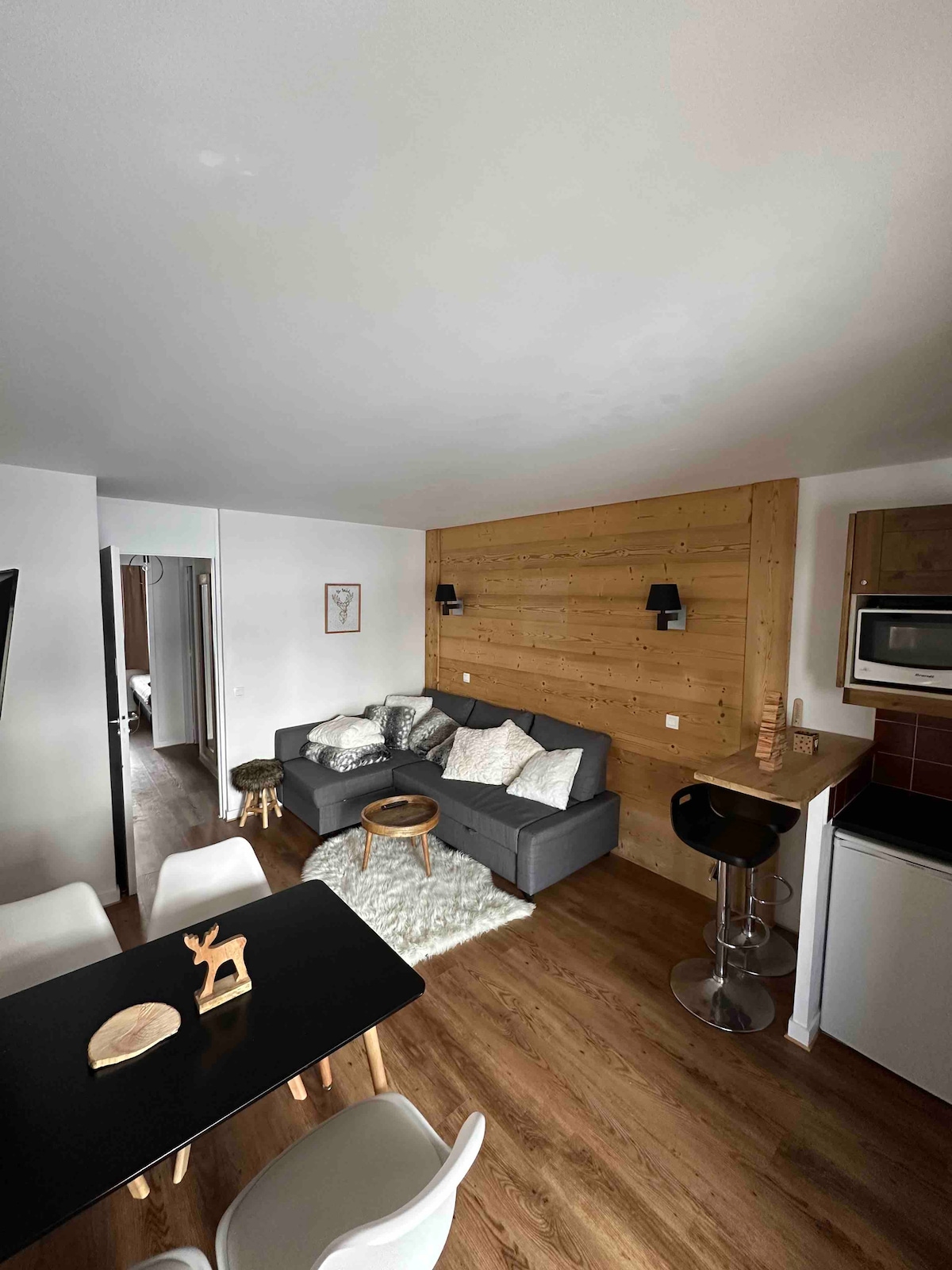 An inviting living area features a gray sofa adorned with several cushions, complemented by a wooden accent wall. A round coffee table is positioned in front of the sofa. A sleek dining table with white chairs can be seen nearby, along with a modern bar stool.