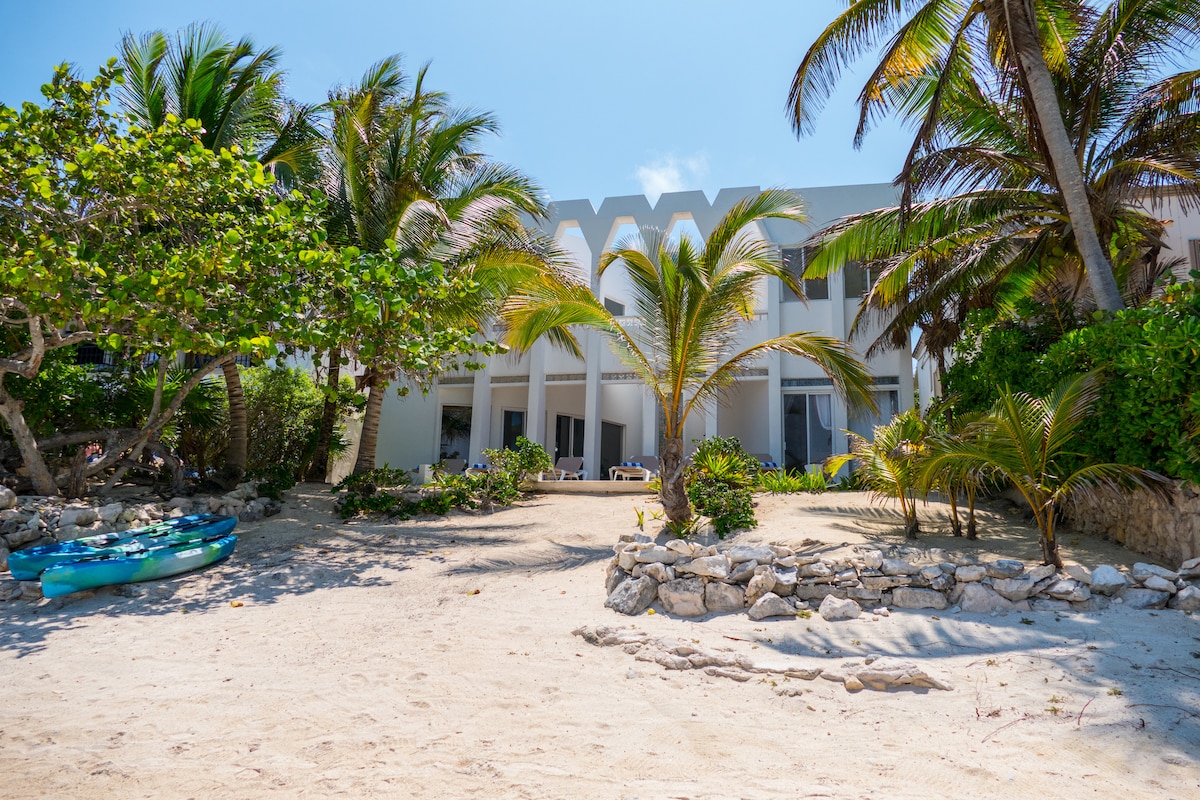 A beachfront view showcases the villa's architecture with Mayan arches, framed by lush palm trees. A sandy area leads to the ocean, where two kayaks are positioned on the shore, inviting guests for water activities.