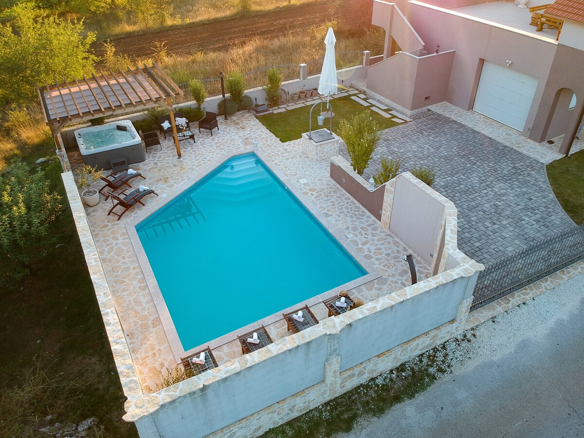 An aerial view of the outdoor area highlights a rectangular pool surrounded by a stone patio. Sun loungers are positioned along the poolside, and a shaded area with seating is visible. A hot tub is situated nearby, with greenery providing a natural backdrop.