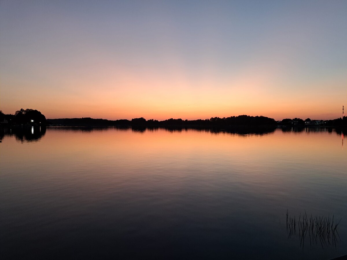A serene lake scene is captured at sunset, with soft hues of orange and purple reflecting on the calm water surface. The horizon is lined with silhouettes of trees, creating a tranquil and peaceful atmosphere.