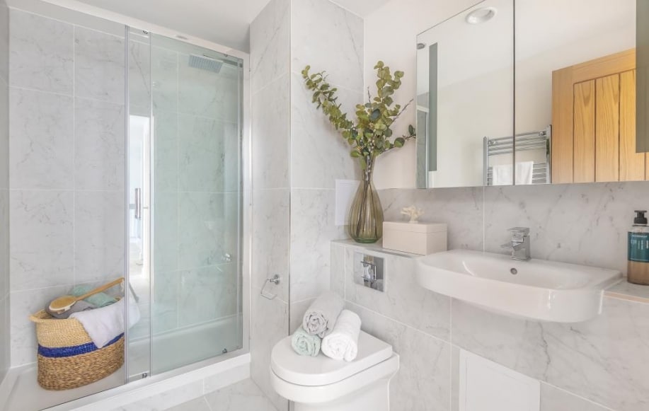 A modern bathroom features a glass shower enclosure and a sleek sink with a mirror above. Natural light reflects off the marble walls. A decorative vase filled with greenery adds a touch of nature, complemented by neatly arranged towels on the countertop.