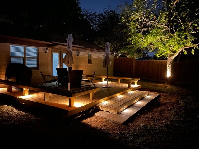 A well-lit wooden deck extends from the back of the house, furnished with two umbrellas and several chairs. Soft lighting outlines the deck, creating a cozy ambiance against the evening sky. A tree stands nearby, adding natural beauty to the outdoor space.