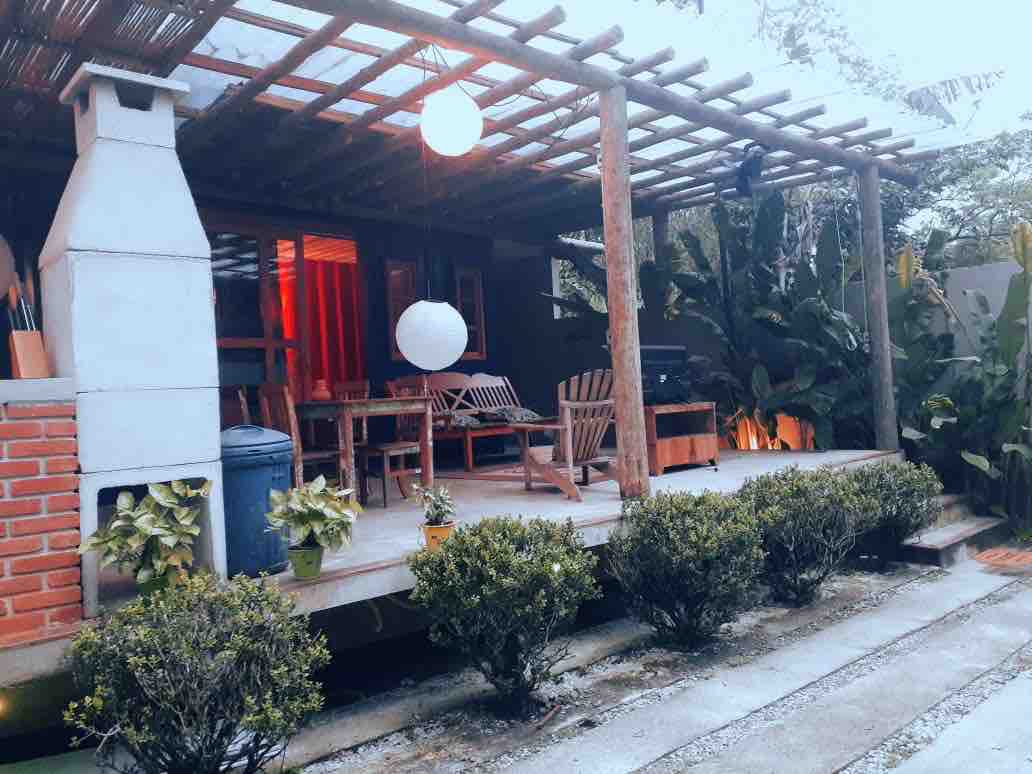 A charming outdoor space features a wooden deck with seating arranged around a central table. Lush greenery frames the area, and ambient lighting creates a serene atmosphere. A stylish lantern hangs above, complementing the rustic elements of the surrounding architecture.