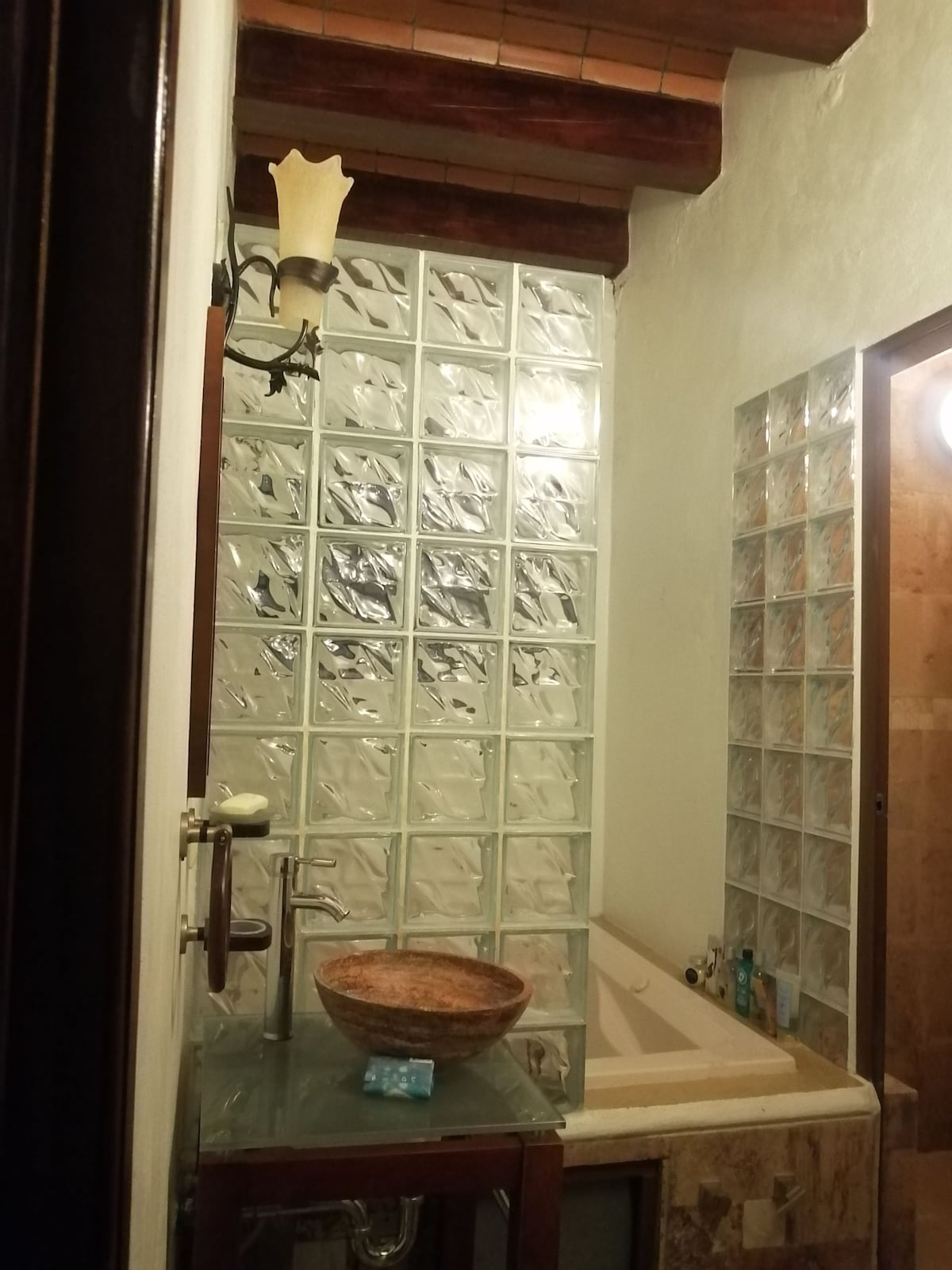 A modern bathroom is presented with glass block walls allowing natural light. A spacious jacuzzi is positioned beside a decorative stone sink. Shelves display toiletries, contributing to an organized and functional space. The warm-tone lighting highlights the textured surfaces and natural materials.