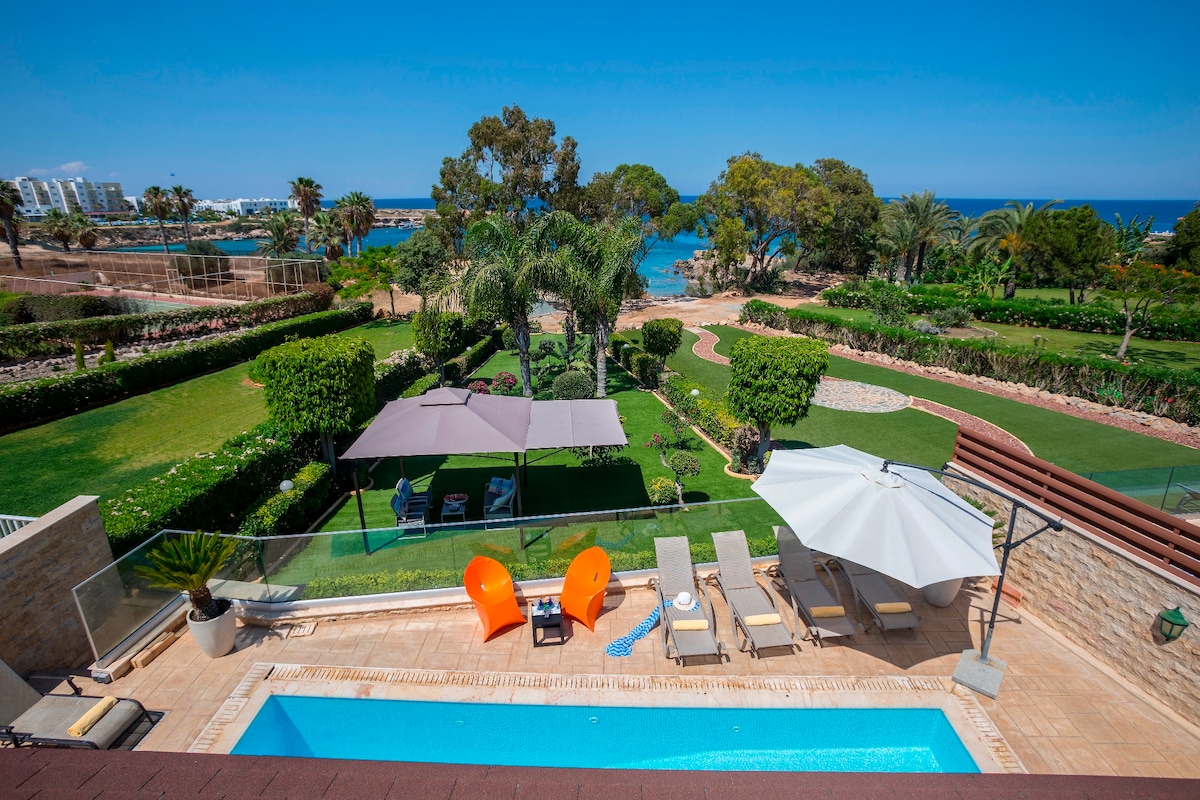 The image showcases a panoramic view of the outdoor area, featuring a private heated pool surrounded by comfortable sun beds. A spacious garden with mature greenery is visible, along with shaded seating areas beneath umbrellas. The distant coastline adds to the tranquil seaside atmosphere.