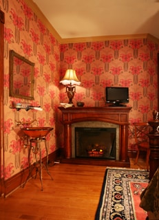 The room features hand-screened wallpaper in rich jewel tones, complemented by a decorative mirror and a decorative lamp. A wooden fireplace, with an electric insert, serves as a cozy focal point. A small table and chair arrangement invites relaxation in this elegantly styled space.