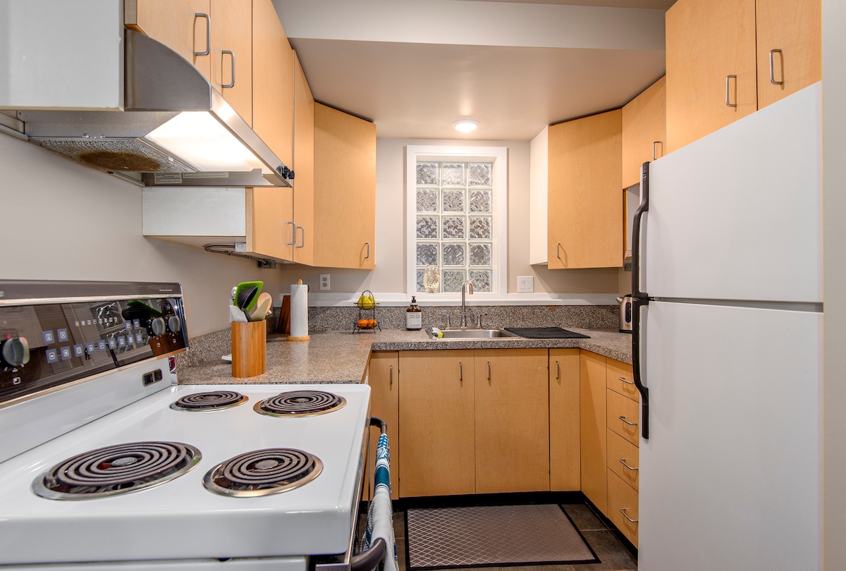 The kitchen features light wood cabinetry and a spacious countertop. Appliances include a white refrigerator and an oven with a stove top. A glass block window provides natural light, while a sink and modern faucet are positioned against the countertop.