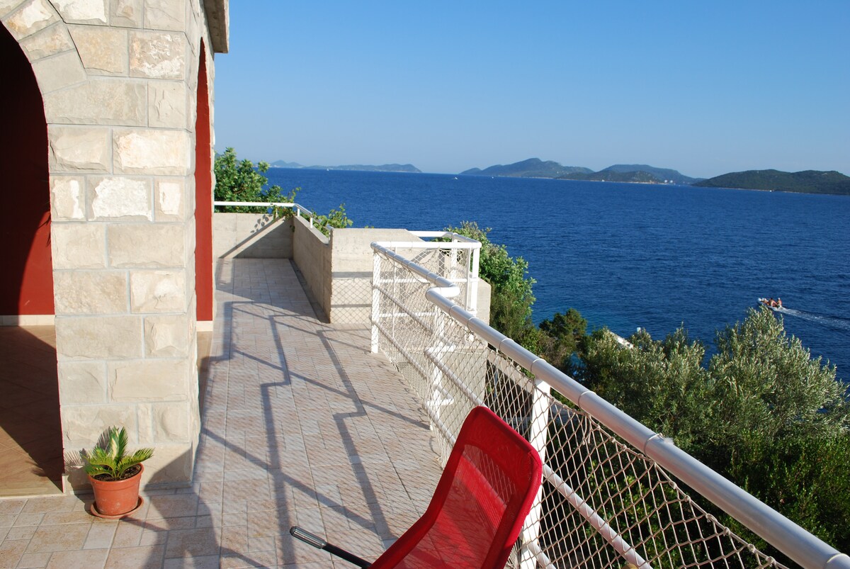A spacious terrace is featured, overlooking the calm blue sea and distant islands. The stone structure adds a rustic charm, while a single red chair is positioned for relaxation. Sunshine illuminates the area, creating a serene outdoor space for enjoyment.