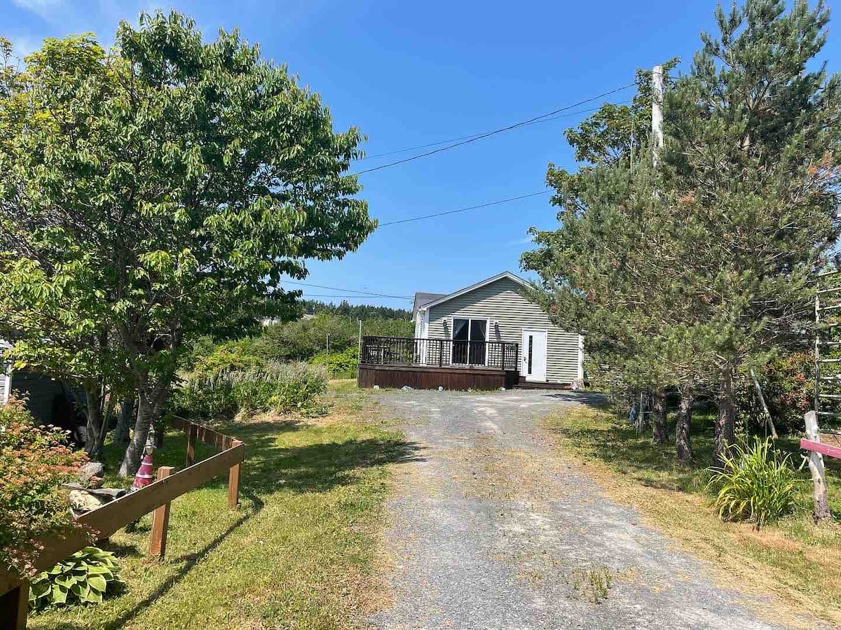 A well-maintained gravel driveway leads up to the cozy home, framed by lush greenery and trees. A wooden deck is visible in front, inviting relaxation with a clear blue sky in the background. The surroundings include a mix of bushes and open grass areas.