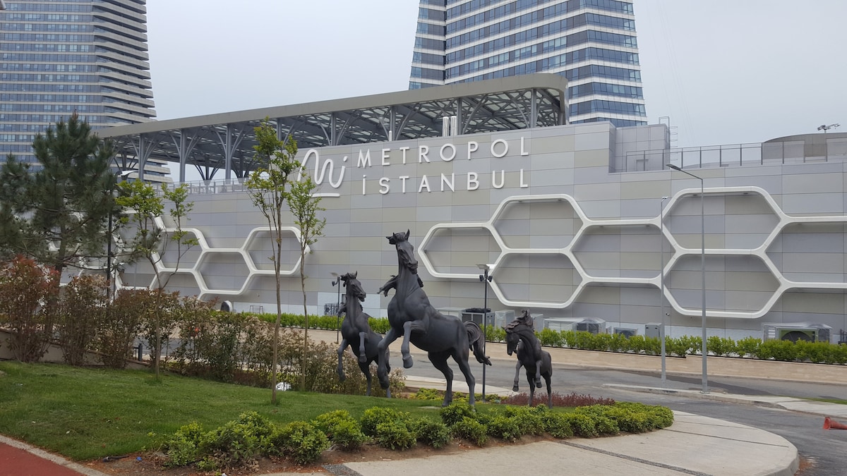 A modern building facade is visible, featuring the name 'Metropol Istanbul' prominently displayed. In the foreground, several bronze sculptures of horses in dynamic poses are positioned along a landscaped area, with greenery and pathways enhancing the urban setting.