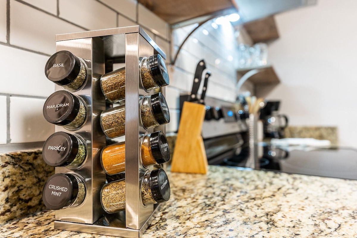 A modern spice rack featuring eight clear spice containers is positioned on a granite countertop. Each container is labeled with its contents, showcasing various herbs and spices. A sleek kitchen setup is visible in the background, including knives on a magnetic strip and glassware on open shelves.