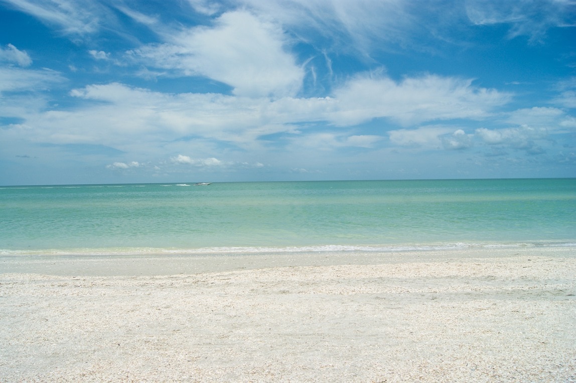 A wide expanse of sandy beach extends toward a calm, aquamarine sea, under a bright sky filled with soft, scattered clouds. The shoreline is lined with small seashells, adding texture to the sand.