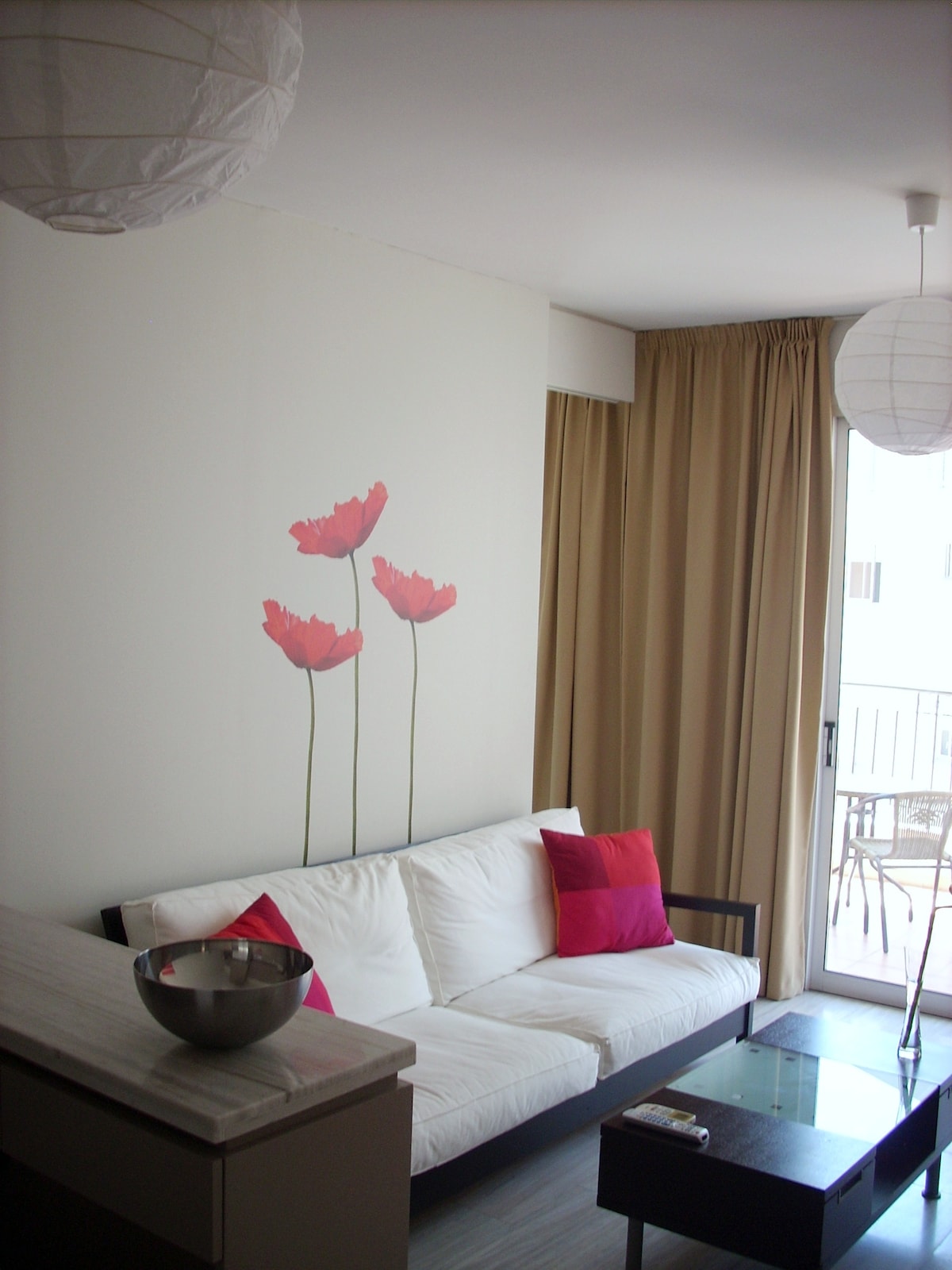 A living room area features a white sofa adorned with colorful cushions. A floral wall decal with red flowers draws attention. Natural light enters through a glass door that leads to a small balcony, complemented by light beige curtains.