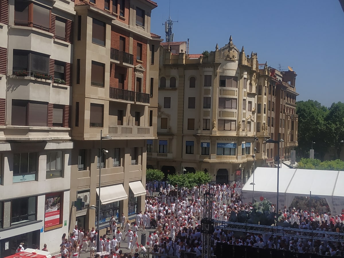A vibrant street scene is captured, showcasing a large crowd dressed in white with red accents. Surrounding historical buildings provide an architectural backdrop under a clear blue sky. Tents and staging areas are visible, indicating an event atmosphere typical of local festivities.