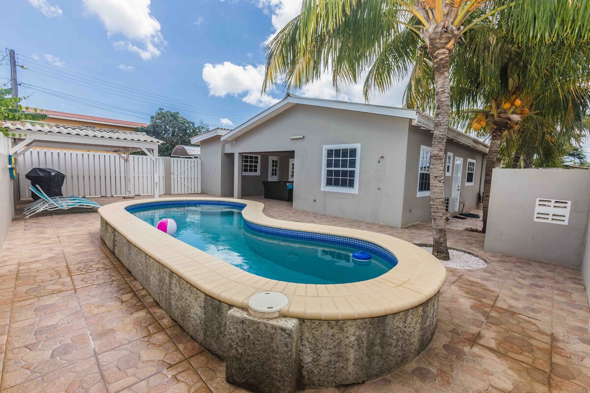 A serene outdoor space features a winding pool surrounded by palm trees. Sunlight reflects off the water's surface, while a comfortable lounge chair sits nearby. The exterior of the house is light-colored with multiple windows, and a private fence adds to the secluded atmosphere.