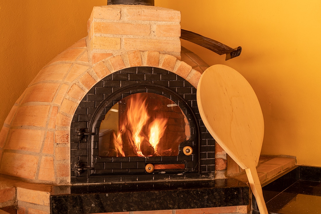 A traditional brick oven is prominently featured, with flames visible inside, emanating warmth. A wooden paddle leans against the oven's exterior. The bright yellow wall serves as a vibrant backdrop, enhancing the rustic charm of the cooking area.