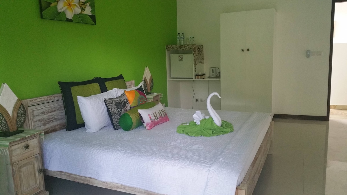 A vibrant bedroom features a light green accent wall, with a spacious bed adorned with decorative pillows and a towel art swan. A compact kitchenette is visible in the corner, alongside a white wardrobe. Natural light brightens the room, enhancing its welcoming ambiance.