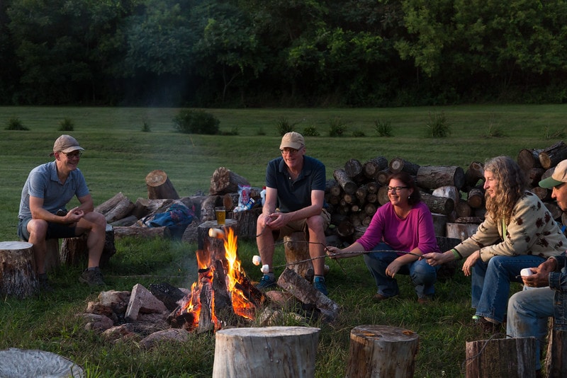 A cozy bonfire scene shows several individuals seated around a crackling fire, roasting marshmallows on sticks. Rustic logs serve as seating alongside stacked firewood, while the surrounding grassy area is edged with trees, creating a peaceful outdoor setting in the background.