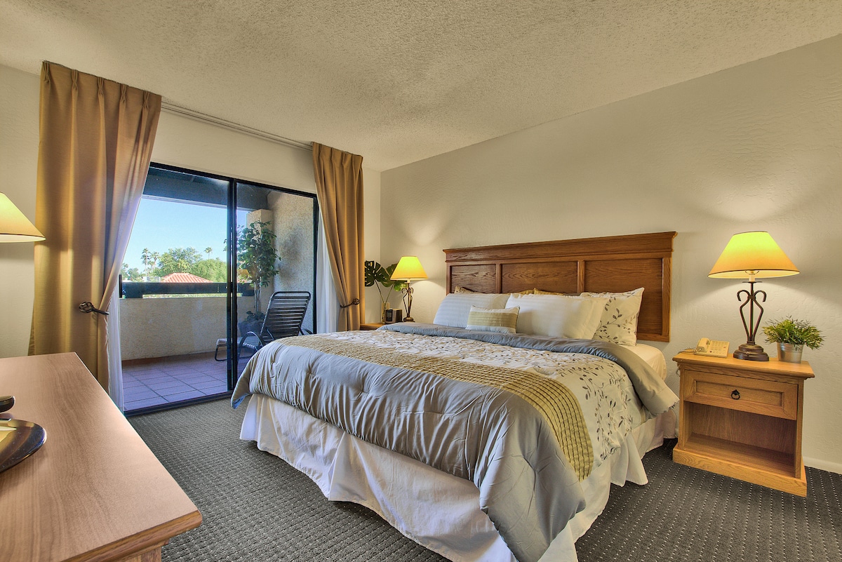 A spacious bedroom features a large bed dressed in neutral linens, complemented by bedside tables with lamps. Soft light filters through the sliding glass door, leading to a private patio. Warm curtains frame the doorway, adding to the inviting atmosphere.