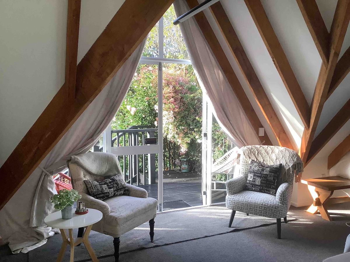 A cozy seating area features two patterned armchairs facing a glass door that opens to a private deck surrounded by greenery. The vaulted ceiling highlights wooden beams, and soft drapes frame the entrance, creating a serene ambiance.