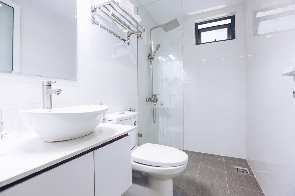A modern bathroom is equipped with a large sink set on a minimalistic countertop. A glass shower stall, with a rainfall showerhead, complements the clean white tiles. A toilet is positioned alongside, with neatly arranged towels hanging on a chrome rack.