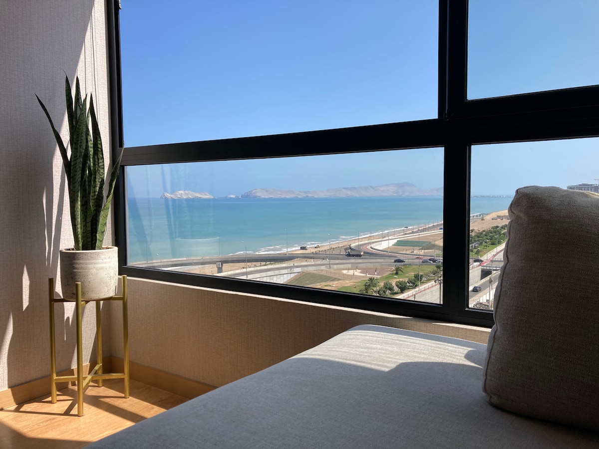 A corner of the room is highlighted by a large window, offering a clear view of the ocean and coastline. A potted plant rests on a gold accent table, complementing the natural light that fills the space.