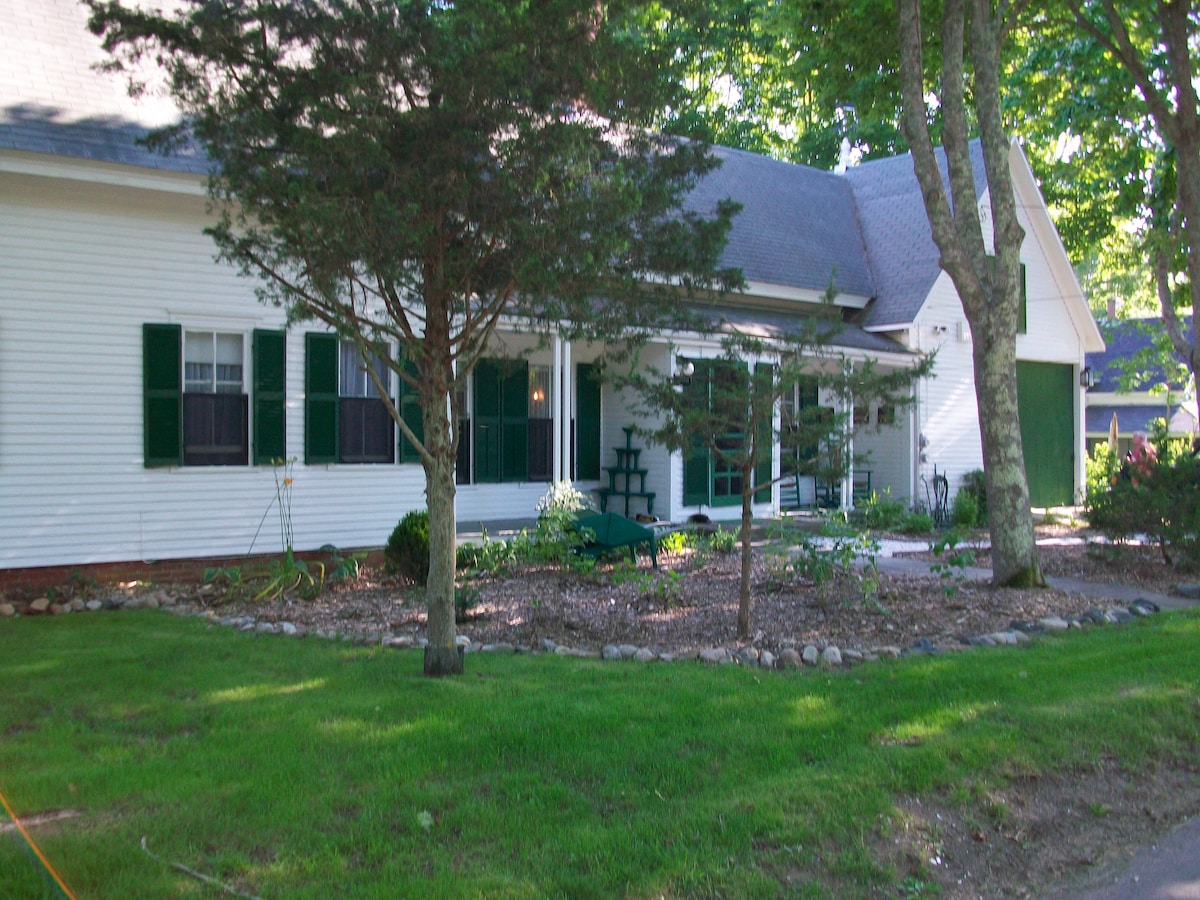 The exterior of the historic home is framed by lush greenery and a well-kept garden. A wide porch with seating is visible, alongside large windows adorned with green shutters. Stone borders enhance the landscaping, and the surrounding lawn is neatly maintained.