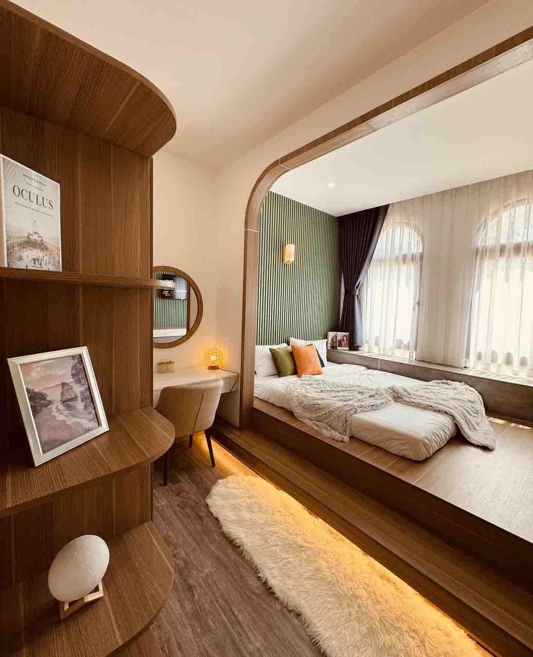 A modern bedroom features a wooden bed platform with an arrangement of neutral bedding. Natural light filters through large windows adorned with sheer curtains. A bookshelf with decorative items, a round mirror above a small desk, and a cozy chair are visible, creating a relaxing atmosphere.